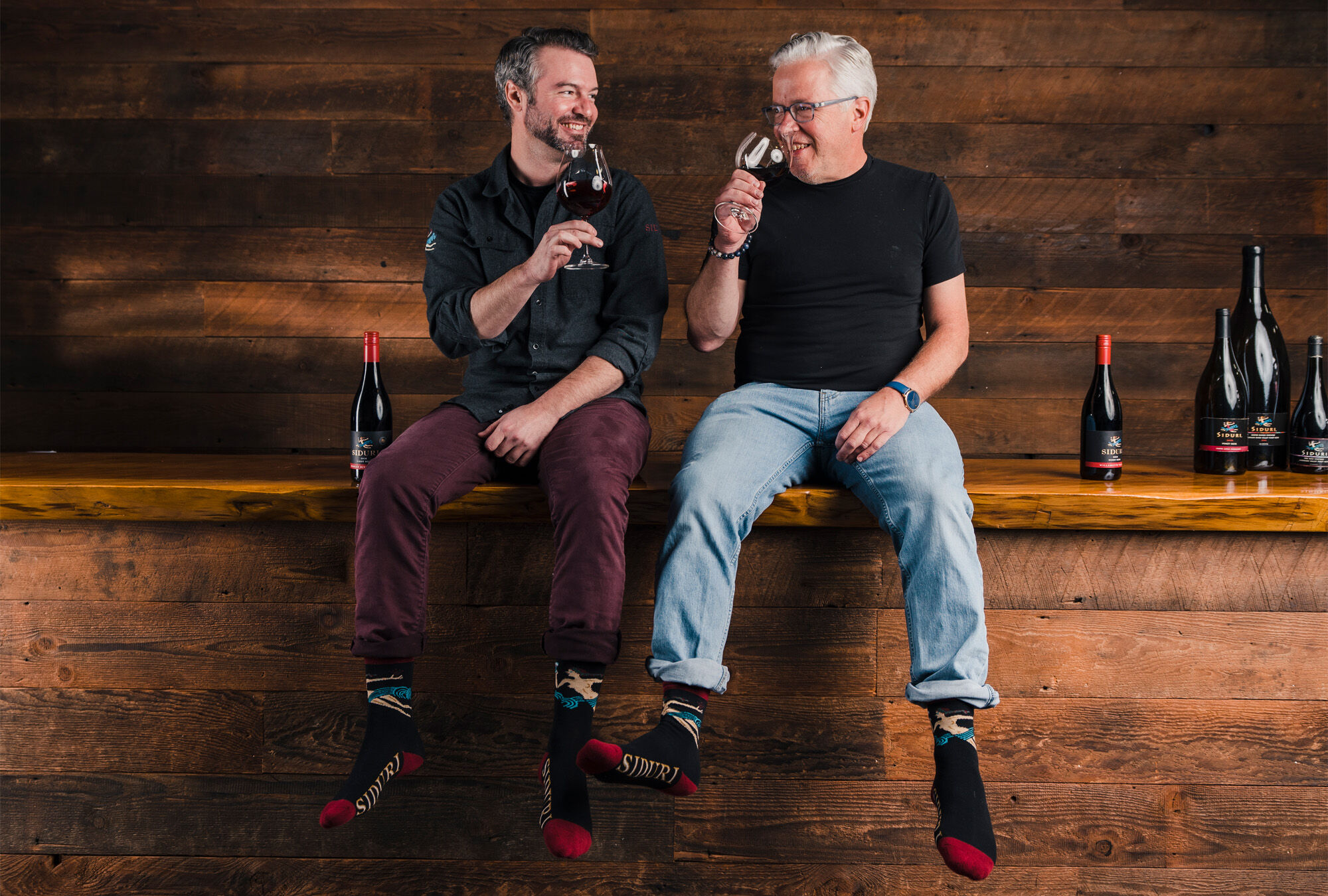 Two men sitting on a wood bench with a matching wood background holding a glass of red wine, with five bottles of red wine on the bench.
