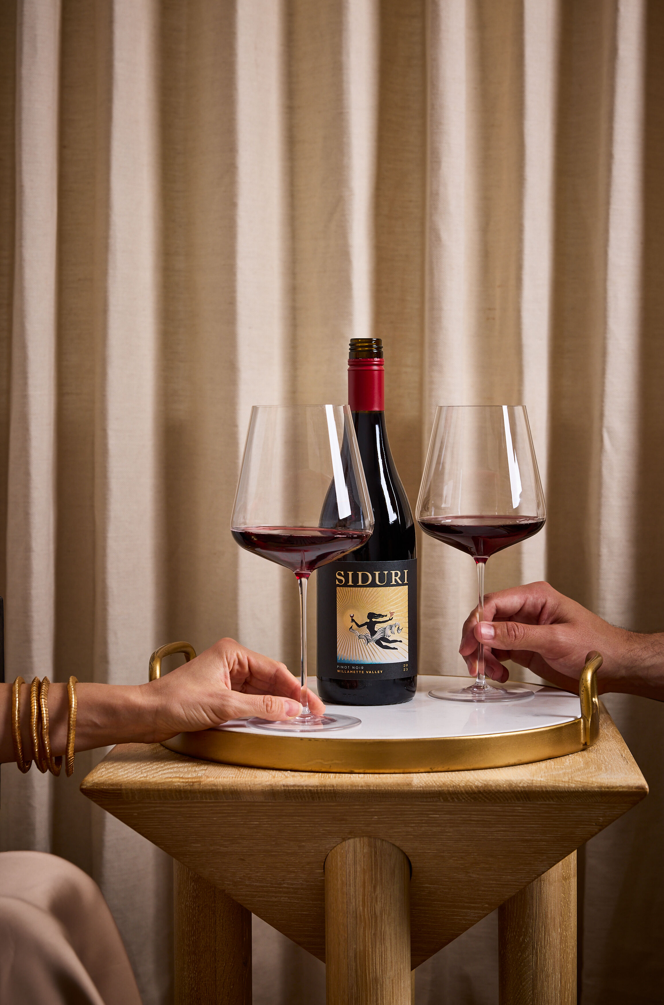 A bottle of red wine on a wood table with beige curtains in the background, and two people's hands holding wine glasses